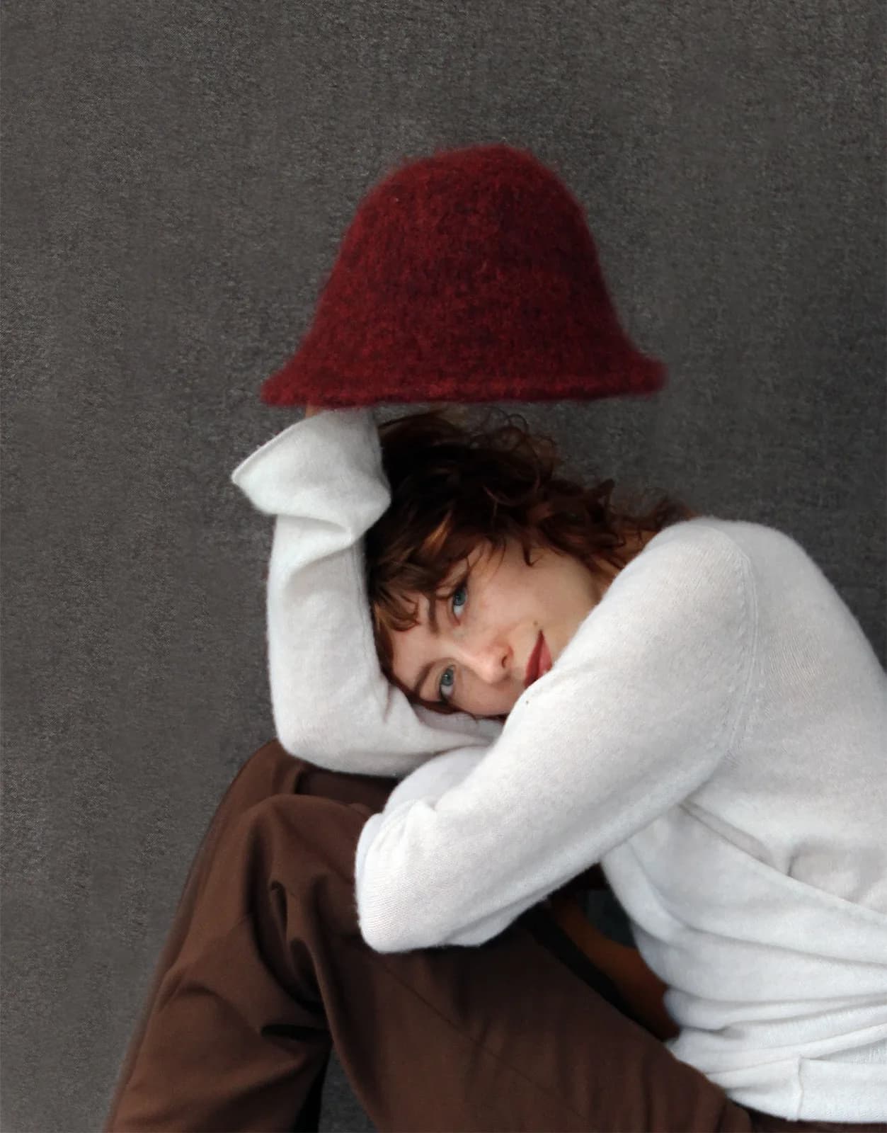 A person with short curly hair resting their head on their arm, balancing a deep red felted bucket hat above them.